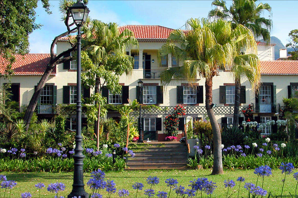 The front façade of the Jardins do Lago hotel with a flower garden and palm trees at the entrance, in Funchal.
